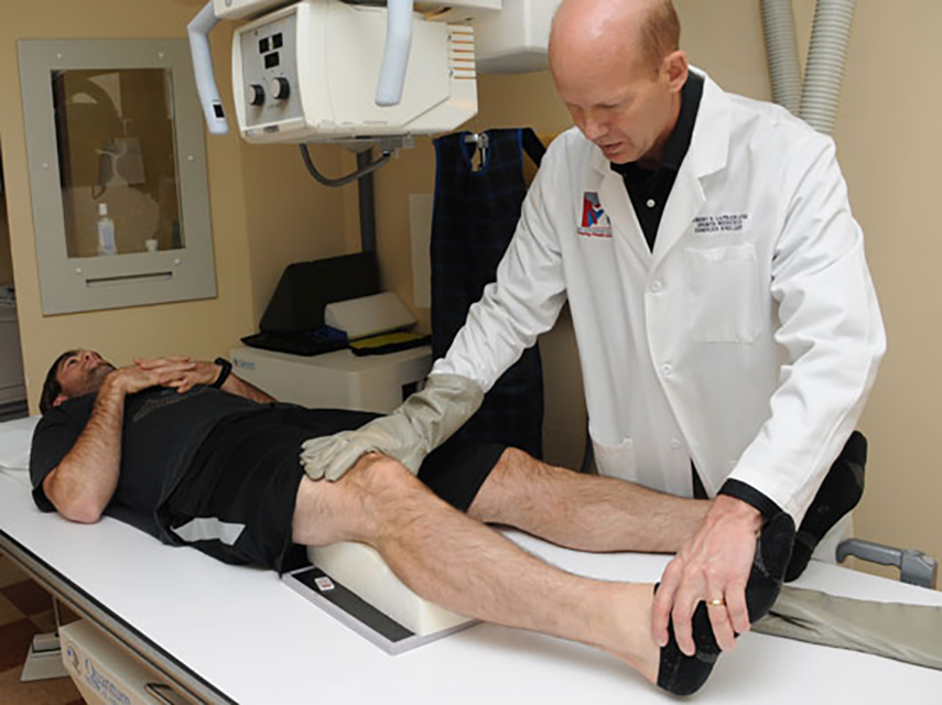 A doctor positioning a man's knee on a foam wedge while the man is lying down on an X-ray table.