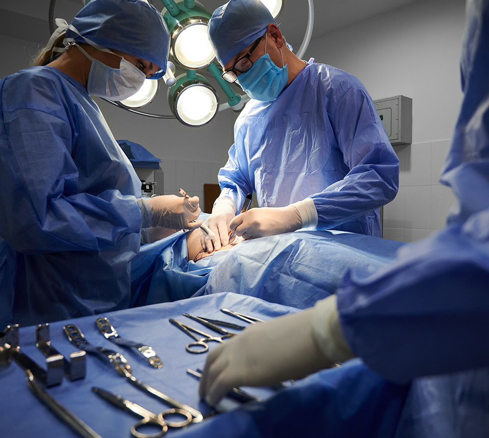 A photo of a person's hand with a glove on reaching for a surgical instrument with surgeons operating on a patient in the background.
